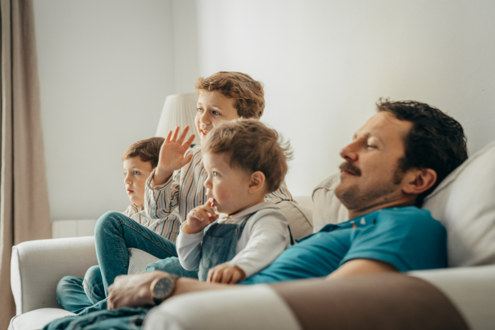 otec unaveně sedí se svými třemi dětmi na gauči a společně se dívají na televizi
