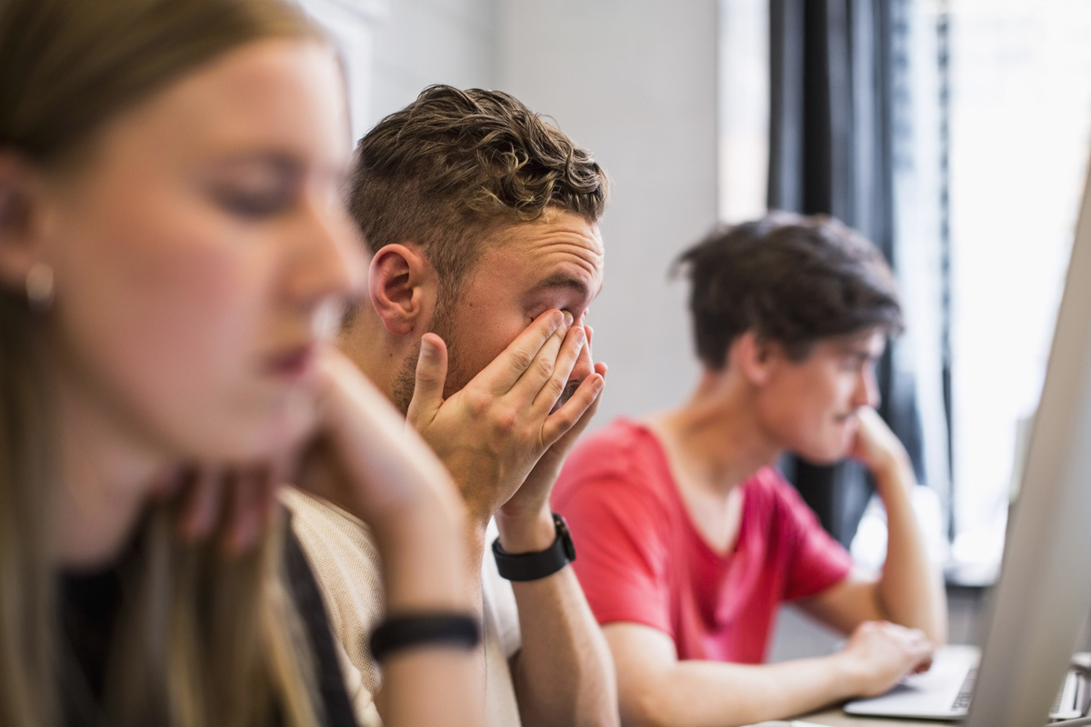 tři studenti sedící na hodině ve škole, přičemž jeden z nich vypadá vystresovaně
